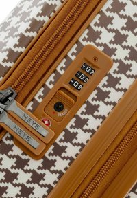 Maleta rígida con estampado de pata de gallo en marrón y blanco, que presenta un cierre de cremallera marrón, candado TSA y una combinación numérica. El tirador de la cremallera tiene la marca.