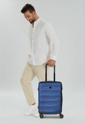 Hombre con camisa blanca y pantalones beige de pie, sonriendo, sosteniendo el asa extendida de una maleta azul con cuatro ruedas.