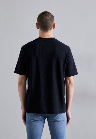 T-shirt preta de manga curta com textura e bainha reta, vestida ao contrário, combinada com jeans azul claro. Design minimalista sem branding visível.