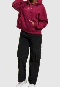 Bordeauxroter Kapuzenpullover mit Kordeln und Fronttasche, kombiniert mit schwarzen Cargohosen mit Seitentaschen und lockerem Schnitt.