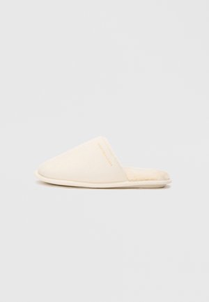 Zapatillas deslizantes de color crema con una textura suave, que presentan una punta redondeada y un sutil acento de logo en dorado en el lateral.