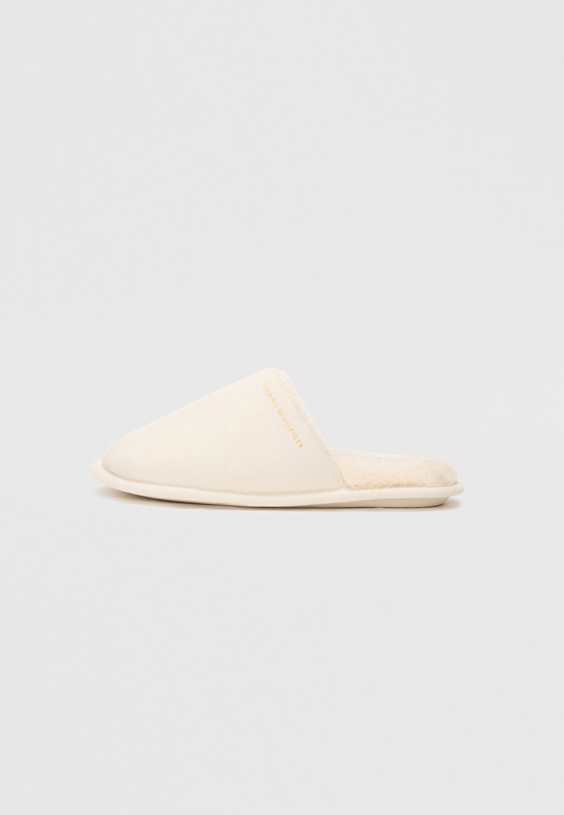 Krém színű, slip-on papucsok, puha textúrával, lekerekített orral és egy visszafogott, arany színű logóval az oldalon.