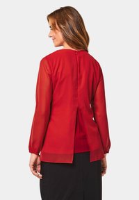 Rode blouse met lange doorschijnende mouwen, een gespleten achterkant en een sluiting bij de hals. Glad materiaal met een getailleerde pasvorm.