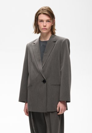 Jeune personne aux cheveux mi-longs portant une veste de costume grise oversize et un pantalon assorti sur un fond blanc uni.