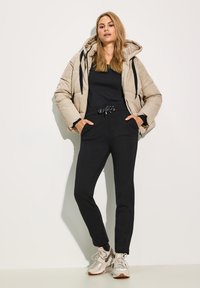 Doudoune beige avec capuche et cordons noirs, associée à un jogging noir et des sneakers gris clair. Textures lisses et design décontracté.