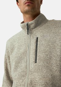 Beige Fleecejacke mit hohem Kragen, Frontreißverschluss und kontrastierenden hellgrauen Akzenten. Verfügt über eine mit Reißverschluss verschließbare Brusttasche und strukturierter Stoff.