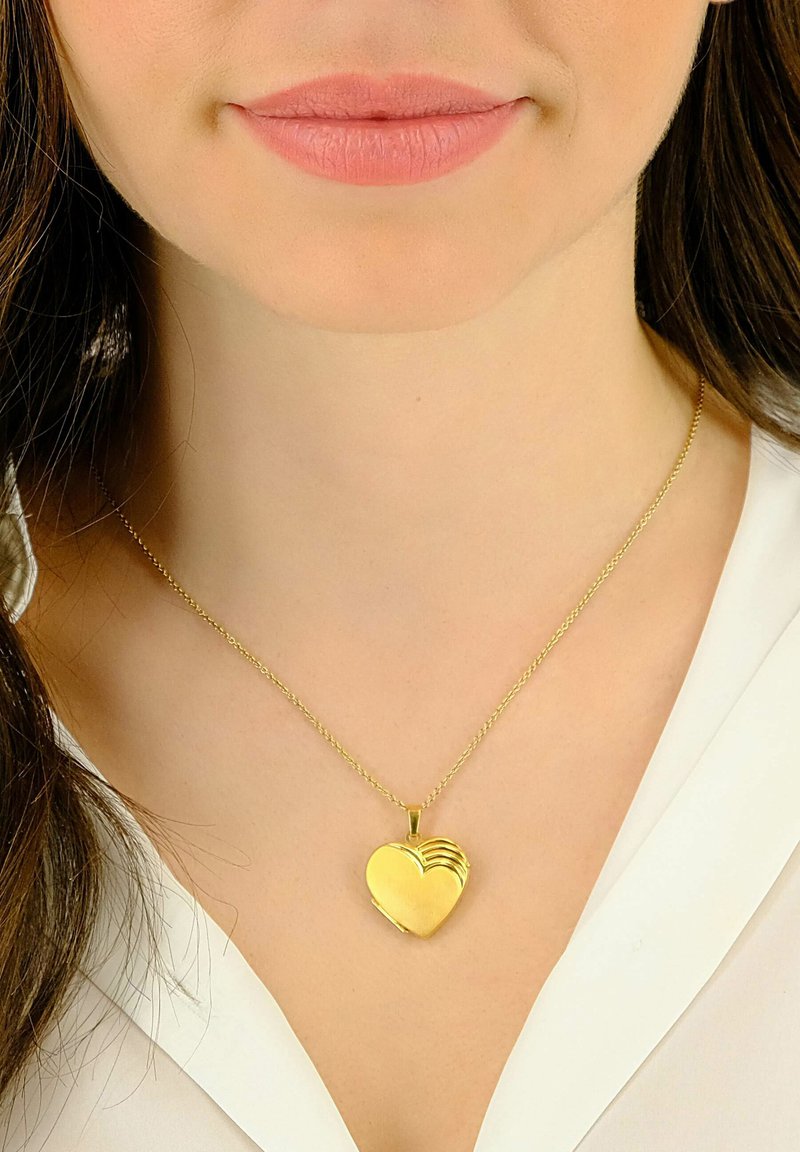 Collier avec pendentif en forme de cœur en or, au fini poli et aux rainures subtiles, suspendu à une délicate chaîne en or, porté contre un chemisier blanc.