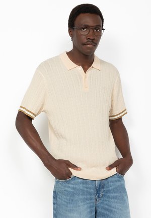 Man met een bril, crème kabelgebreide poloshirt met bruine gestreepte mouwen en blauwe spijkerbroek, poserend met handen in de zakken tegen een witte achtergrond.