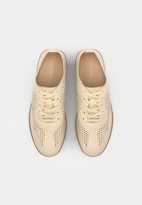 Paire de baskets beiges à lacets avec un design en maille perforée et des semelles marron clair, vues de dessus sur un fond uni.