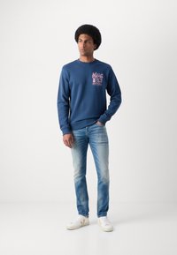 Blaues Sweatshirt mit gerippten Bündchen und Saum, das ein pinkes Grafikdesign auf der linken Brust hat. Kombiniert mit hellblauen Jeans und weißen Sneakers.