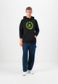 Young man standing wearing black hoodie with yellow circular logo, blue wide-leg jeans, and white sneakers against white background.