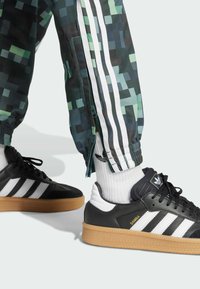 Schwarz-weiße Sneaker mit Gummisohlen, ausgestattet mit einem Design aus drei Streifen. Begleitet von grünen Jogginghosen mit Pixelmuster und weißen Akzenten.