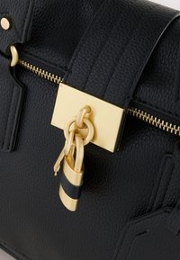 Sac à main en cuir noir texturé avec fermeture éclair dorée, fermoir rectangulaire et un cadenas doré avec clés assorties attachées.