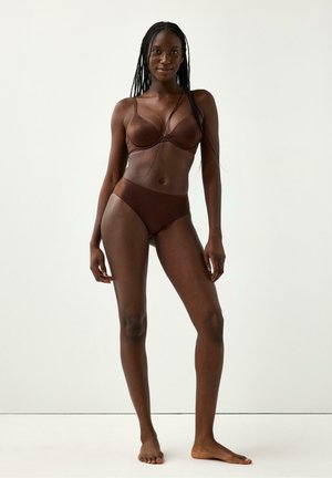 Kvinde med langt flettet hår står barfodet iført brun bh og matchende højtsiddende undertøj mod en ensfarvet hvid baggrund.