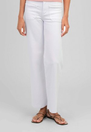 Femme portant un pantalon blanc à jambes larges avec un détail tressé sur les poches avant et des sandales plates marron à lanières, debout sur un sol gris.