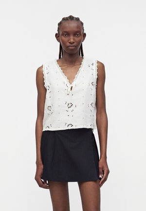 Žena s vrkočmi, ktorá nosí biely bezrukávový top s dierkami a zvlnenými okrajmi a čiernu sukňu, stojaca pred jednoduchým pozadím.