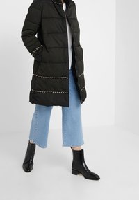 Czarna pikowana kurtka z ozdobnymi złotymi łańcuchami, zestawiona z jasnoniebieskimi jeansami o szerokich nogawkach i czarnymi botkami za kostkę. Gładka faktura i prosta sylwetka.