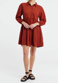 Šatyně v barvě rzi s tečkovaným vzorem, knoflíkovým zapínáním vpředu, límečkem, dlouhými rukávy a řaseným pasem. V kombinaci s černými sandály na pásku.