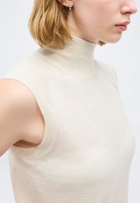 Ξανθιά γυναίκα που φοράει μπεζ αμάνικη μπλούζα ζιβάγκο και μικρό ασημένιο σκουλαρίκι-κρίκο, στραμμένη προς τα δεξιά σε λευκό φόντο.