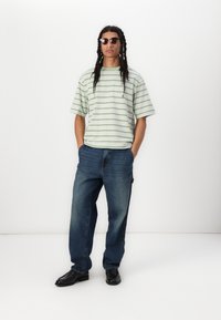 Camiseta de gran tamaño a rayas verdes y blancas, jeans azules de corte holgado, zapatos negros, usando gafas de sol, de pie contra un fondo blanco.