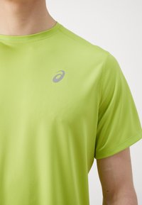 Light green short-sleeve athletic shirt with a round neckline and subtle horizontal texture. Features a gray logo on the upper left chest.