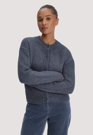 Frau mit zurückgebundenem Haar, die einen dunkelgrauen, geknöpften Strickcardigan und blaue Cordhosen trägt, mit verschränkten Armen.