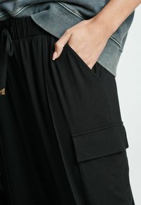 Main dans la poche d'un pantalon cargo noir à rabat, associée à un sweat-shirt gris, montrant la texture du tissu et un style décontracté.