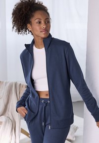 Marineblauwe-ritsjack met hoge kraag, zachte stof en zijzakken. Gecombineerd met een witte crop top en bijpassende marineblauwe sweatpants.