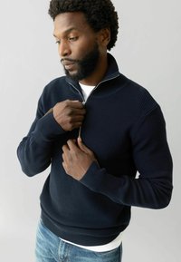 Marineblauer Reißverschluss-Pullover mit geripptem Textur, hohem Kragen und einfarbigem Design. Modell trägt ein weißes T-Shirt darunter und helle Jeans.