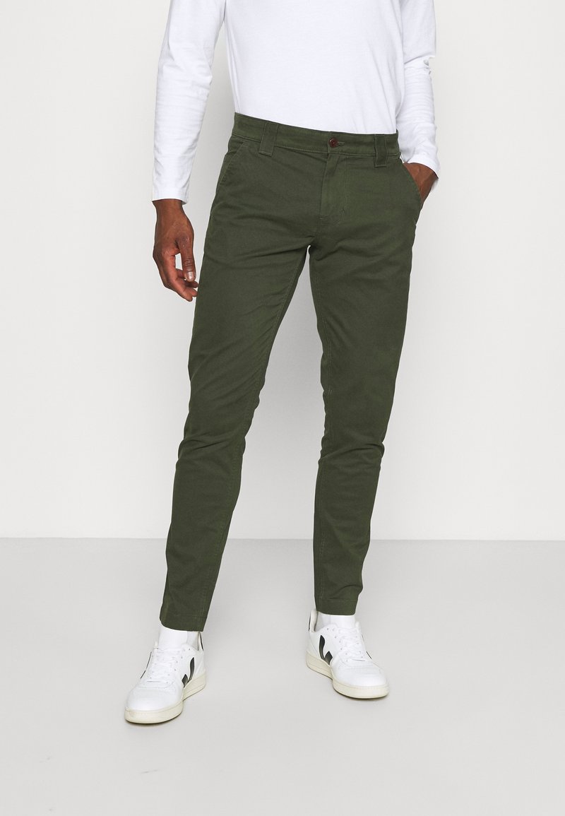 Homme portant un pantalon slim vert olive, une chemise blanche à manches longues et des baskets blanches, debout avec une main dans la poche.
