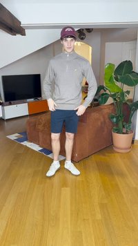 Graues langärmliges Zip-Up-Oberteil, marineblaue Shorts und weiße Turnschuhe. Dunkelrotes Cap. Minimales Design. Innenraum mit einer Couch und Pflanzen.