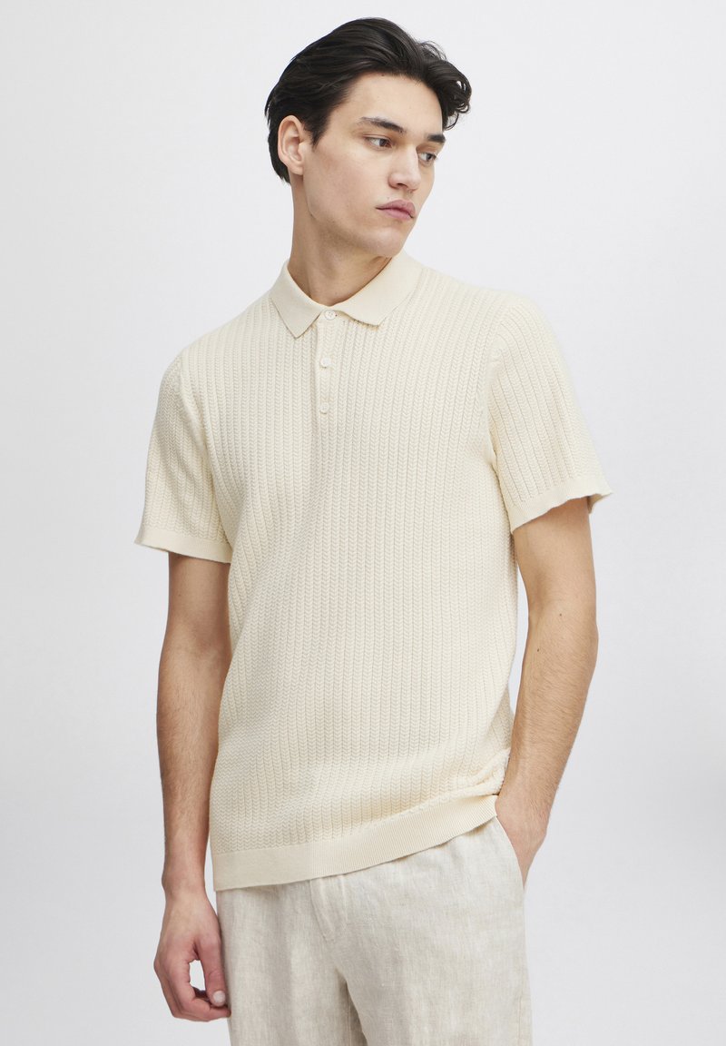 Polo de punto texturizado en crema con mangas cortas y cuello, que presenta una tapeta de tres botones y un corte relajado. Combinado con pantalones de color claro.