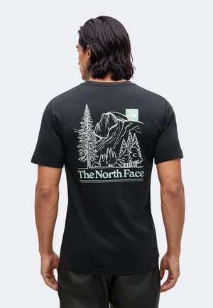 Muž s tmavými vlasmi po ramená nosiace čierne tričko The North Face s bielym a zeleným grafickým motívom hory a stromu na chrbte.
