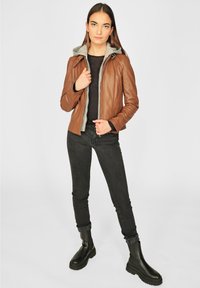Braune Lederjacke mit grauem Kapuzenfutter, Reißverschluss und tailliertem Design. Kombiniert mit schwarzen Jeans und Stiefeletten.