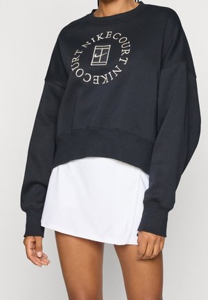 Czarna krótka bluza z napisem "NIKE COURT" i logo w kształcie koła, noszona na białą spódnicę tenisową na stojącej osobie.