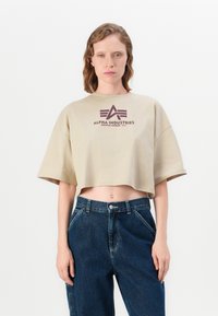 Avkuttet beige T-skjorte med rund hals og korte ermer, med et trykt logo i mørk maroon. Kombinert med høytlivs mørk denim jeans.