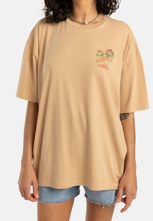 Persona vistiendo una camiseta beige de gran tamaño con un pequeño gráfico de una palmera y el texto "Billabong", combinada con pantalones cortos de mezclilla azul claro.