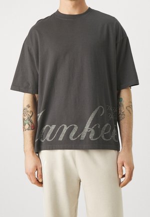 Mężczyzna noszący ciemnoszarą oversize koszulkę z dużym kursywnym napisem "Yankees" i jasne spodnie, pokazujący tatuaże na przedramionach.