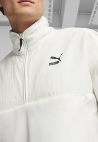 Pullover in pile bianco con colletto alto, chiusura a mezza zip e logo Puma nero sul petto. I pannelli testurizzati creano interesse visivo.