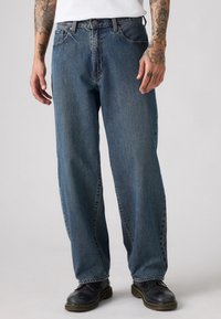 Afslappede fit blå denim jeans, mellemhøj talje, fem-lommydesign, synlig syning, let slidte tekstur, iført sorte sko.