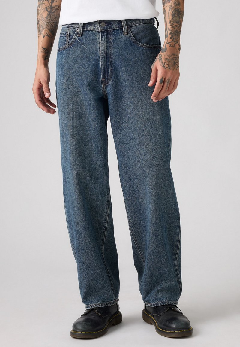 Afslappede fit blå denim jeans, mellemhøj talje, fem-lommydesign, synlig syning, let slidte tekstur, iført sorte sko.