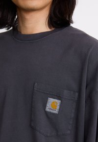 T-shirt à manches longues gris foncé en coton, avec une poche poitrine ornée d'un patch logo jaune et rouge. Texture douce, design à col rond.