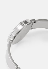 Sølv rustfritt stål klokke med en glatt, polert finish, rund klokkehus, teksturert bezel og en fremtredende krone på siden.