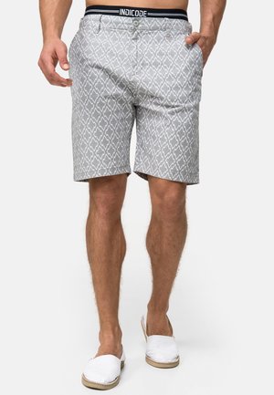 Muž nosící šedé kraťasy s vzorem, s knoflíkem a elastickým páskem s nápisem "INDICODE", v kombinaci s bílými slip-on botami.
