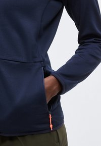 Χέρι στην πλαϊνή τσέπη με φερμουάρ ενός σκούρου μπλε navy μπουφάν, με πορτοκαλί τράβηγμα φερμουάρ, συνδυασμένο με χακί παντελόνι.