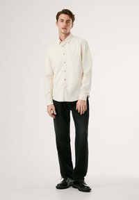 Crème lange mouwen shirt met knoopsluiting en kraag, gecombineerd met donkere spijkerbroek en zwarte veterschoenen, staand op een effen achtergrond.