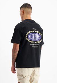 Zwarte T-shirt met een grote grafische afbeelding van een blauwe ovaal omringd door lauwerbladeren en witte tekst. Katoenen materiaal, ontspannen pasvorm.