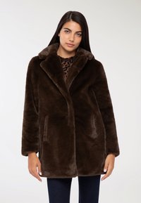 Brązowy futrzany płaszcz z szalowym kołnierzem, dwoma bocznymi kieszeniami i prostym krojem, noszony na ciemnej wzorzystej bluzce i dżinsach.