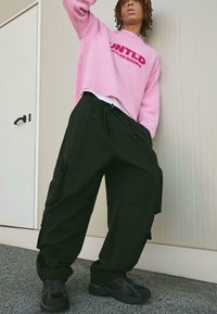 Rózsaszín, túlságosan nagy pulóver "UNTITLD" felirattal, fekete cargo nadrág, amelynek derékrésze zsinórral állítható, és sötét sneakers egy semleges háttér előtt.