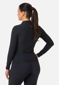 Svart långärmad atletisk topp med ribbad textur, dragkedjeförsedd bakficka och logotypdetalj vid halsringningen. Matchas med svarta leggings.